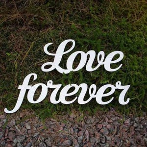 May include: White wooden sign with the words "Love forever" in a cursive font. The sign is placed on a bed of green moss and small rocks, creating a natural backdrop. Ideal for wedding decor.