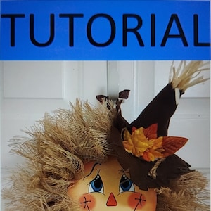May include: A brown and tan scarecrow wreath with a painted face, blue eyes, and a red nose. The scarecrow is wearing a brown hat with fall leaves and a blue and yellow sunflower ribbon.