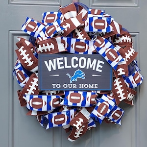 May include: A circular wreath with brown and white football ribbon and blue accents. A rectangular sign in the center reads "WELCOME TO OUR HOME" with a blue lion logo. The wreath is likely for home decor.