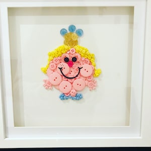 Puede incluir: Una obra de arte enmarcada que presenta a una niña hecha de botones. La niña tiene el pelo amarillo, la cara rosa y zapatos azules. La obra de arte está sobre un fondo blanco.