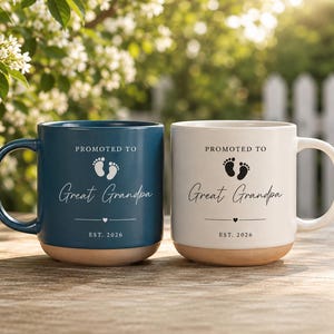Peut inclure: Deux tasses en céramique, une bleu foncé et une blanche, portant l'inscription PROMOTED TO Great Grandpa EST. 2026, une paire d'empreintes de pieds de bébé et un petit cœur. Les deux tasses ont une finition lisse et une base naturelle non émaillée.