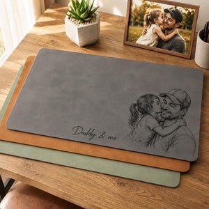May include: Three rectangular desk mats in gray, tan, and green are stacked on a wooden desk. The top gray mat features a sketch of a father and child with the text "Daddy & me". A framed photo and a potted succulent are also on the desk.