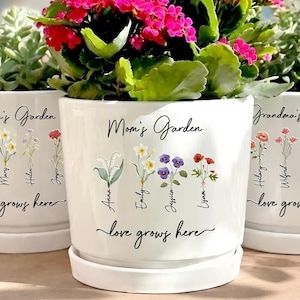 May include: Three white ceramic flower pots with floral designs and the words "Nana's Garden", "Mom's Garden", and "Grandma's Garden". Each pot contains different plants and flowers, including succulents and vibrant pink blooms. The phrase "love grows here" is written on each pot.