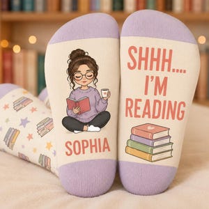 May include: A pair of cream-coloured socks with lavender cuffs. One sock features an illustration of a person with brown hair in a bun wearing glasses and a lavender sweater, reading a book, with the name SOPHIA below. The other says SHHH.... I'M READING above a stack of four colourful books.