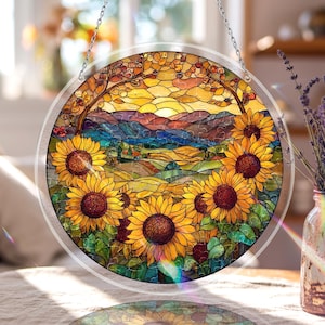 Peut inclure: Vitrail rond représentant un champ de tournesols et un paysage de coucher de soleil. L'œuvre d'art est encadrée d'une bordure transparente et suspendue à une chaîne argentée.