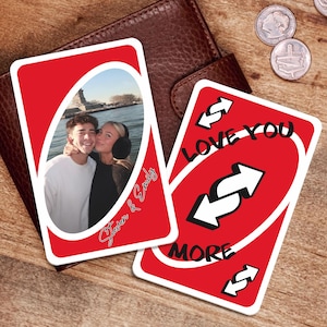 以下が含まれることがあります： 2つのカスタム赤いUnoカード。1枚のカードには「Steven & Emily」というテキストが入ったカップルの写真が写っています。もう1枚のカードには、逆矢印で「Love You More」という言葉が書かれています。カードは茶色の革の財布の上にあります。