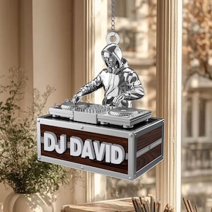 May include: A silver-coloured ornament depicting a DJ in a hooded sweatshirt, operating turntables. The DJ figure is above a wooden box with the text "DJ DAVID" in white letters. The ornament hangs from a silver chain.