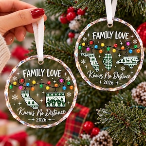 May include: Two round, clear, faceted acrylic Christmas ornaments hanging by white ribbons. Each features the text 'FAMILY LOVE Knows No Distance 2026', a string of colourful holiday lights, snowflakes, and various state map silhouettes.