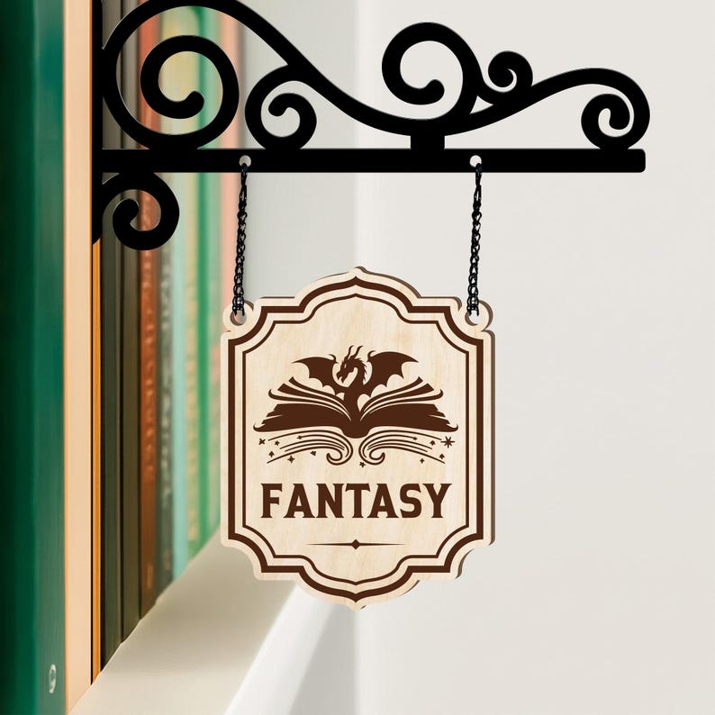 Puede incluir: Un letrero de madera con la palabra "FANTASY" y un drag&oacute;n sobre un dise&ntilde;o de libro abierto, colgado de un soporte de metal negro. El letrero est&aacute; suspendido por dos cadenas y est&aacute; montado en una pared blanca.