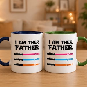 Peut inclure: Deux mugs en c&eacute;ramique blanche avec le texte "I AM THER FATHER" et des graphiques de sabre laser. Un mug a une anse et un bord bleus, l'autre est vert. Chaque mug porte les noms : Emma, James et Emily.