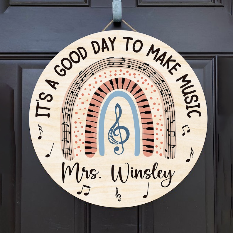 Pu&ograve; includere: Un cartello rotondo in legno con un design a arcobaleno con note musicali e una chiave di violino. Il testo "It's a good day to make music" &egrave; scritto intorno all'arcobaleno. Il nome "Mrs. Winsley" &egrave; scritto sotto l'arcobaleno.