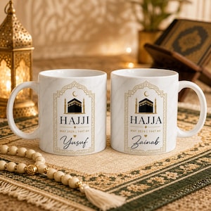 Op de afbeelding: Twee witte keramische mokken met een marmerpatroon, elk voorzien van een goudomlijnd boogontwerp met een illustratie van de Kaaba, een halve maan en de tekst "HAJJI" of "HAJJA" met "MAY 2026 | 1447 AH" en de namen "Yusuf" of "Zainab".