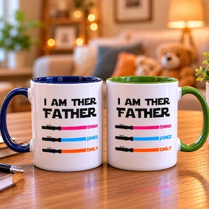 Peut inclure: Deux mugs en c&eacute;ramique blanche avec le texte "I AM THER FATHER" et trois motifs de sabre laser color&eacute;s. Les tasses ont des poign&eacute;es et des bords bleus et verts. Les noms Emma, James et Emily sont imprim&eacute;s en dessous.