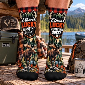 May include: A pair of crew socks featuring a red and black buffalo plaid pattern at the top, with the text Ethan's LUCKY HUNTING SOCKS printed above a graphic of a moose standing in a forest landscape with mountains and a full moon.