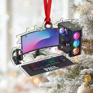 May include: A clear acrylic Christmas ornament shaped like a gaming setup, including a monitor, computer tower, keyboard, mouse, and headset. The ornament is decorated with colorful lights and a red ribbon for hanging. The name "PAUL" is printed on the mousepad.