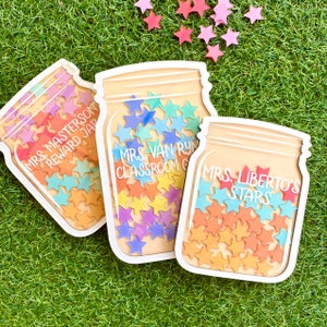 Pode incluir: Três placas de madeira em forma de frasco de pedreiro com adesivos de estrelas coloridas no interior. As placas são etiquetadas como "Mrs. Masterson Reward Jar", "Mrs. Vanryn Classroom Goals" e "Mrs. Liberto's Stars".