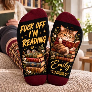 May include: Black socks with maroon cuffs featuring a graphic of a cat wearing glasses reading a book while giving the middle finger. The text reads FUCK OFF I'M READING and Emily. The design includes stacks of books, flowers, and hearts.