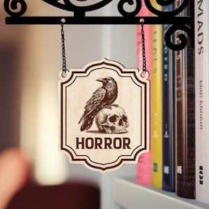 Puede incluir: Letrero de madera con un cuervo posado sobre una calavera, con la palabra "HORROR" en negrita. El letrero est&aacute; colgado de un soporte de metal negro con un dise&ntilde;o decorativo. El fondo incluye una estanter&iacute;a con libros coloridos.