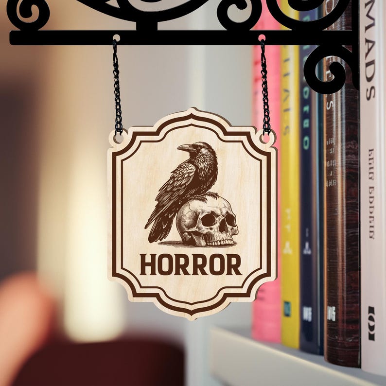 Puede incluir: Letrero de madera con un cuervo posado sobre una calavera, con la palabra "HORROR" en negrita. El letrero est&aacute; colgado de un soporte de metal negro con un dise&ntilde;o decorativo. El fondo incluye una estanter&iacute;a con libros coloridos.