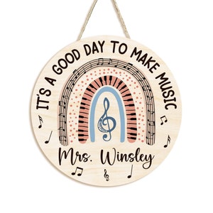 Pu&ograve; includere: Un cartello rotondo in legno con un design a arcobaleno che presenta note musicali e una tastiera di pianoforte. Il cartello recita "It's a good day to make music" e "Mrs. Winsley".