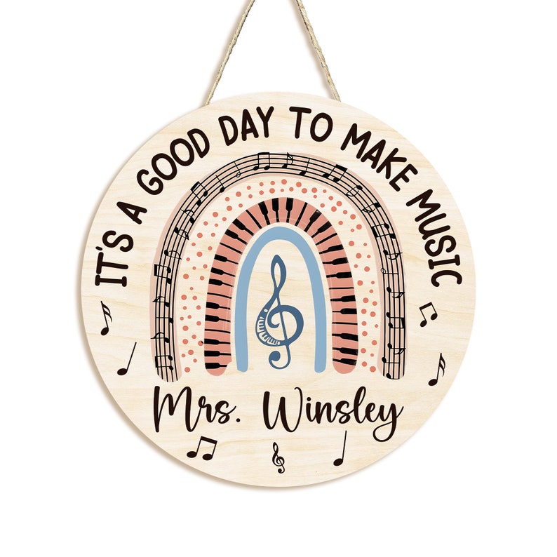 Pu&ograve; includere: Un cartello rotondo in legno con un design a arcobaleno che presenta note musicali e una tastiera di pianoforte. Il cartello recita "It's a good day to make music" e "Mrs. Winsley".