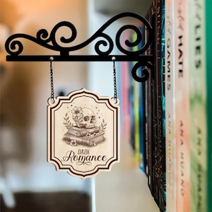 Puede incluir: Un letrero de madera con las palabras "Dark Romance" y una ilustraci&oacute;n de calavera, colgado de un soporte de metal negro. El letrero presenta una calavera sobre libros y flores. El soporte est&aacute; fijado a una estanter&iacute;a llena de libros.