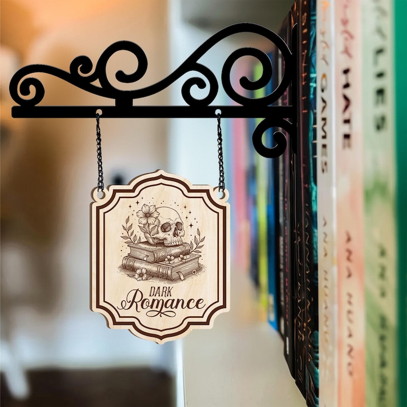 Puede incluir: Un letrero de madera con las palabras "Dark Romance" y una ilustraci&oacute;n de calavera, colgado de un soporte de metal negro. El letrero presenta una calavera sobre libros y flores. El soporte est&aacute; fijado a una estanter&iacute;a llena de libros.