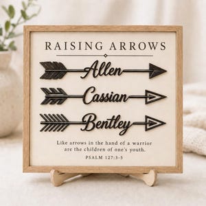Puede incluir: Un letrero cuadrado de madera con fondo claro que presenta el texto RAISING ARROWS en la parte superior. Tres iconos de flechas negras se muestran horizontalmente, cada uno incorporando los nombres Allen, Cassian y Bentley en cursiva.