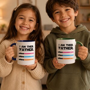 Peut inclure: Deux mugs en c&eacute;ramique blanche avec le texte "I AM THER FATHER" et des noms personnalis&eacute;s. Un mug a une anse et un int&eacute;rieur bleus, l'autre a une anse et un int&eacute;rieur verts. Les mugs ont un design de sabre laser color&eacute;.