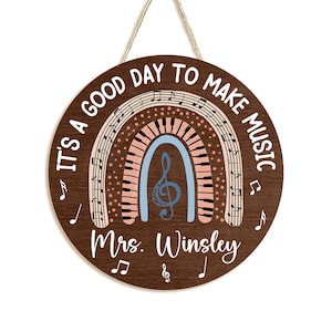 Pu&ograve; includere: Un cartello rotondo in legno marrone con il testo "It's a good day to make music" in lettere bianche. Un arcobaleno con note musicali e tasti di pianoforte si trova al centro del cartello. Il testo "Mrs. Winsley" &egrave; in fondo al cartello.