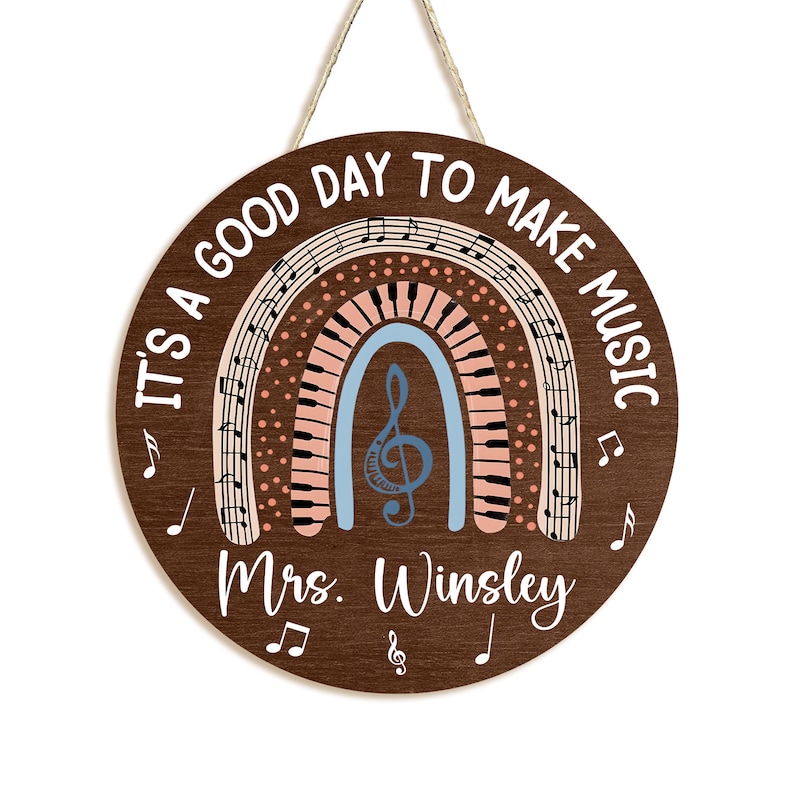 Pu&ograve; includere: Un cartello rotondo in legno marrone con il testo "It's a good day to make music" in lettere bianche. Un arcobaleno con note musicali e tasti di pianoforte si trova al centro del cartello. Il testo "Mrs. Winsley" &egrave; in fondo al cartello.