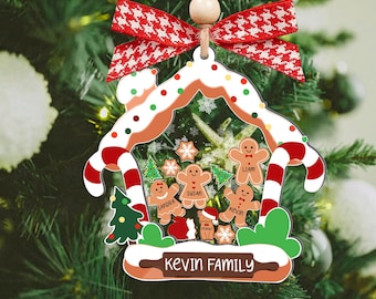 Puede incluir: Adorno navideño con forma de casa de jengibre, decorado con bastones de caramelo y hombrecitos de jengibre. El adorno incluye el texto "KEVIN FAMILY" y está rematado con un lazo a cuadros rojos y blancos.