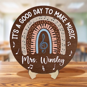 Pu&ograve; includere: Un cerchio di legno marrone con il testo "It's a good day to make music" attorno al bordo. Un arcobaleno con note musicali e tasti di pianoforte &egrave; al centro. Il testo "Mrs. Winsley" &egrave; sotto l'arcobaleno.