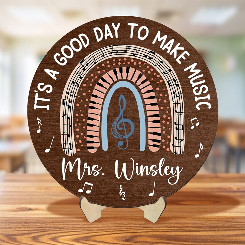Pu&ograve; includere: Un cerchio di legno marrone con il testo "It's a good day to make music" attorno al bordo. Un arcobaleno con note musicali e tasti di pianoforte &egrave; al centro. Il testo "Mrs. Winsley" &egrave; sotto l'arcobaleno.