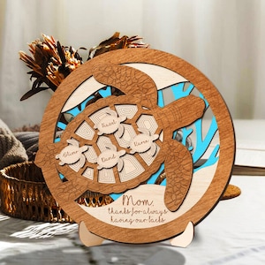 May include: A round wooden plaque featuring a laser-cut sea turtle design. The plaque has a brown frame with a light blue background and includes the names Oliver, David, Hazel, and Kara. The text reads, "Mom, thanks for always having our backs."