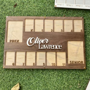 May include: A wooden photo frame with 12 slots for pictures, labeled with grades from Pre-K to Senior. The frame is personalized with the name "Oliver Lawrence".