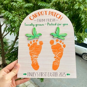 Op de afbeelding: Een houten bord met een wortelveldthema. Het heeft twee oranje voetafdrukken met groene bladeren, de tekst "CARROT PATCH FARM FRESH Locally grown - Picked for you" en "FINLEY'S FIRST EASTER 2025".