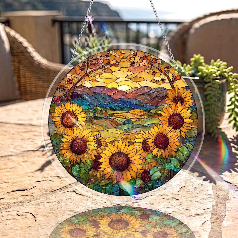 Op de afbeelding: Een rond gebrandschilderd glazen raam met een zonnebloemontwerp. Het kunstwerk toont een landschap met zonnebloemen, glooiende heuvels en een zonsondergang. Het glas is omlijst met een heldere rand en hangt aan een zilveren ketting.