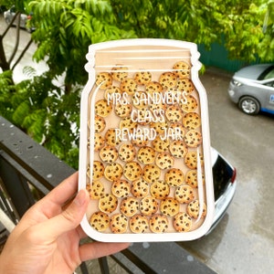 以下が含まれることがあります： チョコレートチップクッキーで満たされた、白い瓶型の報酬チャート。「MRS. SANDVEN'S CLASS REWARD JAR」というテキストが白で表示されています。瓶は、緑の葉と車の背景の前に保持されています。