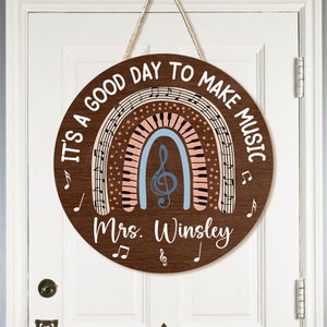 Pu&ograve; includere: Un cartello rotondo in legno con un design a arcobaleno che presenta note musicali e una chiave di violino. Il cartello recita "It's a good day to make music" e "Mrs. Winsley".