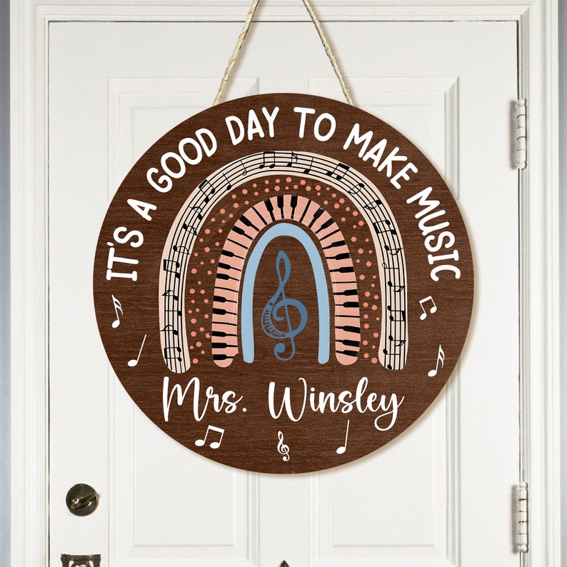 Pu&ograve; includere: Un cartello rotondo in legno con un design a arcobaleno che presenta note musicali e una chiave di violino. Il cartello recita "It's a good day to make music" e "Mrs. Winsley".