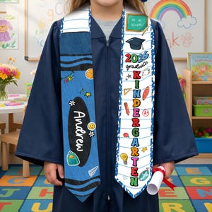 May include: A navy blue graduation gown with two personalized stoles. One stole is blue with the name "Andrew" and school-themed graphics. The other stole is white with "2026 graduate Kindergarten" text and school icons.