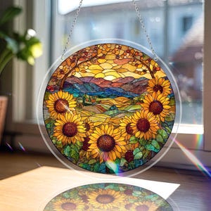 Op de afbeelding: Een rond gebrandschilderd raam met een levendig zonnebloemveld en een zonsonderganglandschap. De zonnebloemen hebben gele bloemblaadjes en donkerbruine centra. De achtergrond toont glooiende heuvels, bergen en een kleurrijke lucht. Het raam heeft een heldere rand en hangt aan een zilveren ketting.