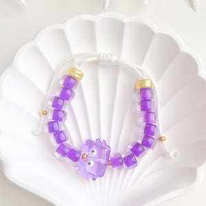 May include: A bracelet featuring translucent purple square beads and a central purple cat-shaped charm. The beads are strung on a white cord with gold-toned cylindrical spacers and small gold-toned beads, finished with an adjustable sliding knot closure.