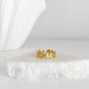 May include: A gold-toned ring featuring a repeating pattern of small, textured seashell shapes arranged in a continuous band. The ring has an open design, allowing for slight adjustments to the fit, and displays a polished, reflective metallic finish.