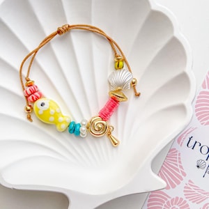 May include: A colorful beaded bracelet with a yellow fish charm, pink and white beads, and a gold-colored clasp. The bracelet is displayed on a white seashell-shaped dish. A card with the text "tropical vibe" is in the background.