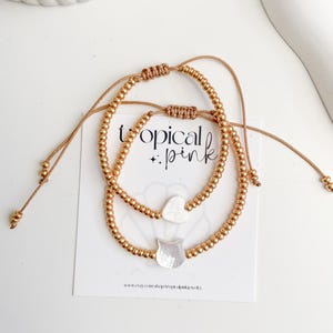 May include: Two gold beaded bracelets with adjustable cords. Each bracelet features a small, white, cat-shaped charm and a heart-shaped charm. The bracelets are displayed on a white card with the text "tropical pink" and a website address.