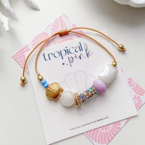 May include: A gold-toned beaded bracelet with a seashell charm, blue and white beads, and a central section of iridescent rhinestones. The bracelet is on a card with the text "tropical + pink".