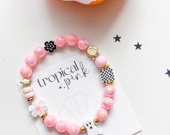 Könnte beinhalten: Ein Perlenarmband mit rosa facettierten Perlen, einem weißen Geisteranhänger, einem schwarz-weiß karierten Würfel, einer weißen Blume und goldenen Sternakzenten. Das Armband liegt auf einer weißen Karte mit dem schwarzen Aufdruck tropical + pink.
