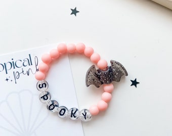 Könnte beinhalten: Ein elastisches Armband mit matten, hellrosa runden Perlen, transparenten Perlen mit schwarzen Buchstaben, die SPOOKY buchstabieren, und einem glitzernden, durchscheinenden, dunkelgrauen fledermausförmigen Anhänger auf einer weißen Tropical Pink Karte.